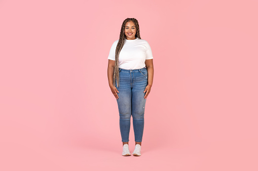 Happy Black Plus Size Female Model Posing In Studio Over Pink Background, Full Length Shot. Braided Lady Standing Smiling To Camera Wearing T-Shirt And Jeans