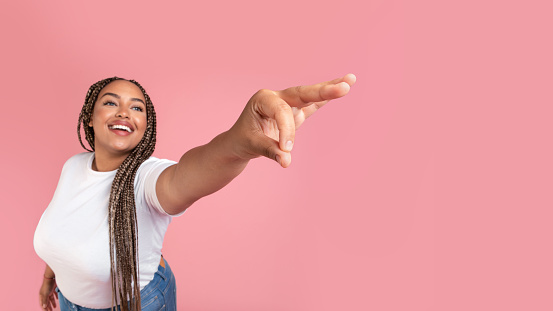 Cheerful African American Overweight Lady Holding Invisible Object In Fingers Showing And Advertising Something Standing Over Pink Studio Background. Panorama, Selective Focus