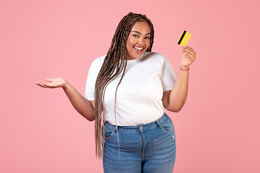 Positive Black Female Holding Credit Card Advertising Bank Offer Posing Smiling To Camera Standing Over Pink Background In Studio. Finances And Great Payment Service Concept