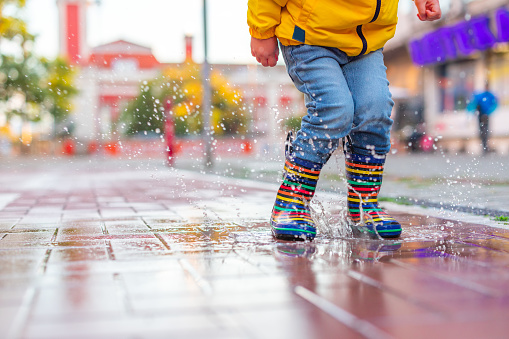Happy little kid boy jumping on rainy puddle in city street, autumn season