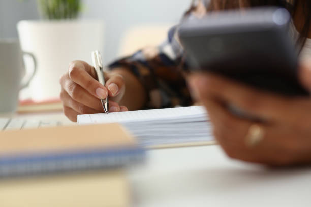 une femme écrit dans un cahier avec un stylo au bureau et tient un smartphone - transportation form photos et images de collection