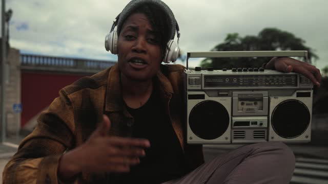 African girl having fun singing while listening to music with headphones and vintage boombox stereo in the city street