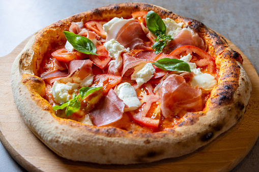 Close-up on round pizza with abundant delicious garnish of prosciutto, mozzarella, tomato slices and basil leaves, served on wooden board