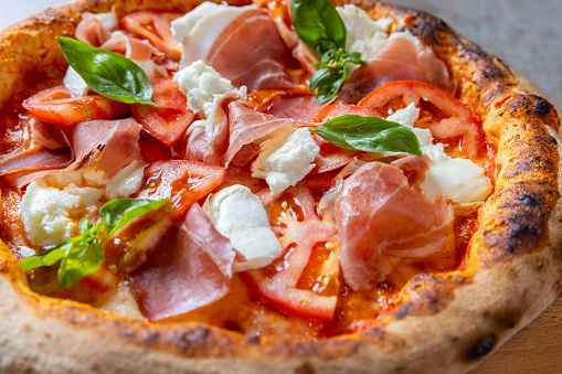 Close-up on round pizza with abundant delicious garnish of prosciutto, mozzarella, tomato slices and basil leaves