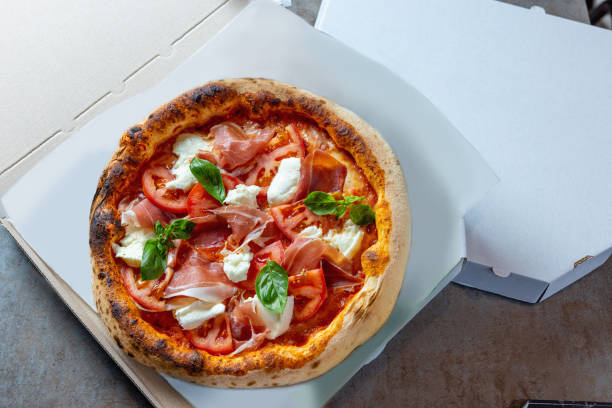 istock Pizza with prosciutto, tomato and mozzarella topping in open box for delivery 1473906582