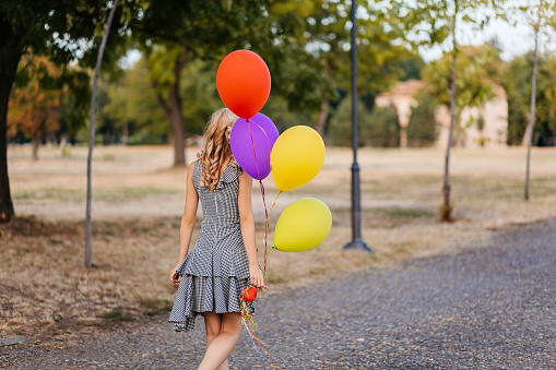 Rear-view of a beautiful young woman holding colorful helium balloons in the public park.