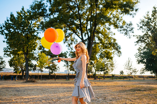 Beautiful young woman holding colorful helium balloons in the public park.