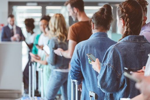 Travelers with luggage using smart phones while waiting in line for boarding at airport. Focus on foreground.