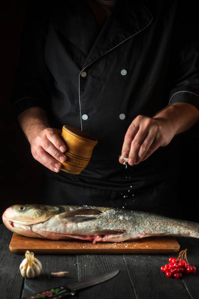 The chef prepares silver carp herring in the restaurant kitchen. The cook sprinkles salt on the fish. Work environment on the kitchen table