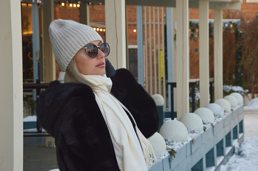 Stylish, fashionable young woman in a white hat, dark fur coat and sunglasses outdoors in the city in cold weather. Beautiful modern girl