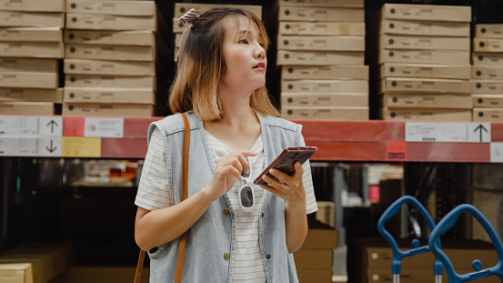 Young Asian woman customer shopping using smartphone searching goods stock in retail warehouse. Business warehouse shopping concept.