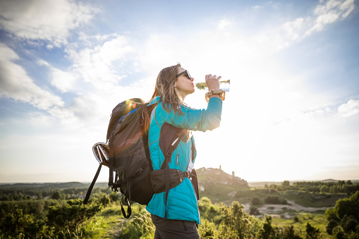 Side view of young woman wearing sports clothes drinking water from a water bottle in the mountain at sunset.