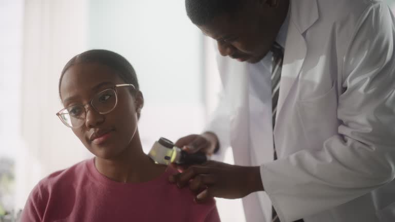 African American Dermatologist Using a Dermatoscope to Identify Worrying Cancerogenic Tissues on the Skin of a Beautiful Young Female During a Health Check Visit to a Clinic. Black Doctor in Hospital