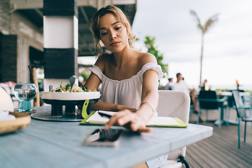 Young ethnic female in casual wear sitting at cafe terrace having lunch and browsing smartphone in tropical place on blurred background