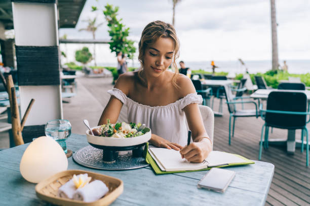 Pensive young ethnic tourist having lunch in summer cafe and taking notes in planner