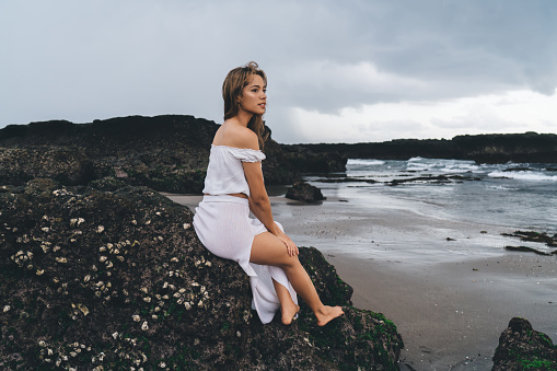 Side view of full body barefoot slim female in white dress sitting on stone cliff and looking away thoughtfully while enjoying vacation