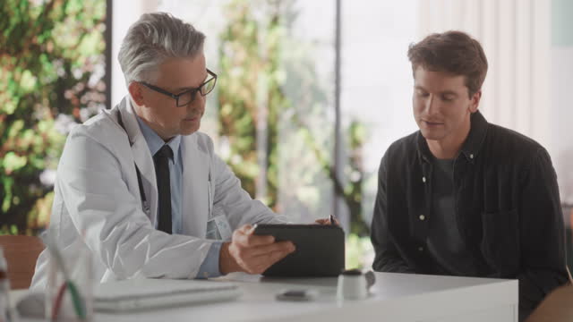 Doctor with Stylish Gray Hair Showing Analysis Results on Tablet Computer to a Healthy Young Man During Visit to a Health Clinic. Physician Working Behind a Desk in Hospital Office