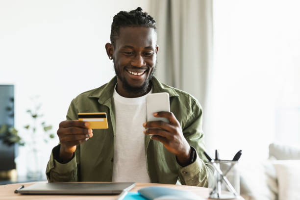 istock Excited black guy using cellphone and holding credit card, shopping online or banking while staying home 1473924158