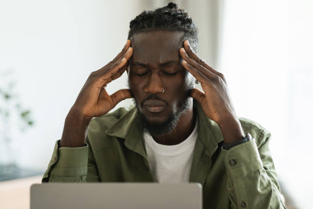 Unhappy black male freelancer touching head, suffering from headache or having issues with work, sitting at desk