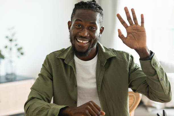 istock Greeting concept. Friendly black man waving at camera and smiling, saying hi while having video call at home 1473924210