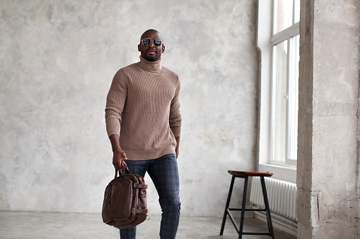 Handsome african american man walking indoors wearing fashionable clothes beige sweater, trousers and leather bag, sunglasses