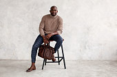 Stylish man sitting on chair and looking at camera. Confident fashion model in fashionable outfit turtleneck knitted sweater, plaid trousers, leather shoes and bag. Office clothes style for busines