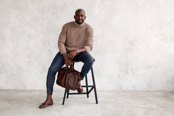 Stylish man sitting on chair and looking at camera. Confident fashion model in fashionable outfit turtleneck knitted sweater, plaid trousers, leather shoes and bag. Office clothes style for busines Stylish man sitting on chair and looking at camera. Confident fashion model in fashionable outfit turtleneck knitted sweater, plaid trousers, leather shoes and bag. Office clothes style cardigan sweater stock pictures, royalty-free photos & images