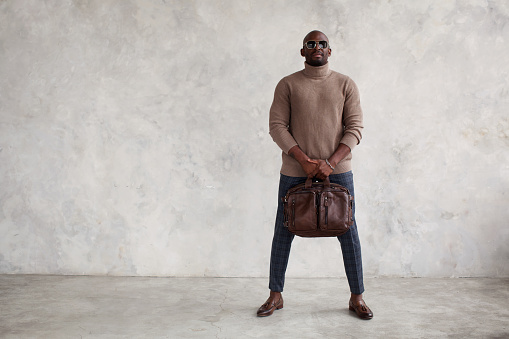 Fashion man stands against gray concrete background, wearing stylish clothes, turtleneck sweater, plaid trousers, brown boots loafers, bag and glasses