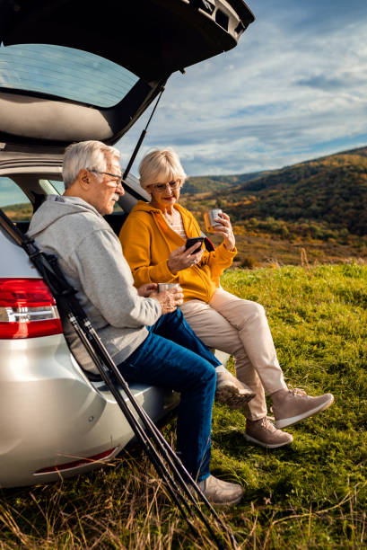 senior couple sitting against the car, resting after hiking in countryside takes photo with smart phone. - old men car vertical imagens e fotografias de stock