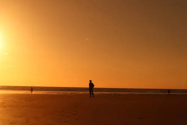 Ambient orange sunset making a silhouette of a man standing on the beach alone with a bright orange sky and waves and sea and coast in background, thoughtful meaningful pose