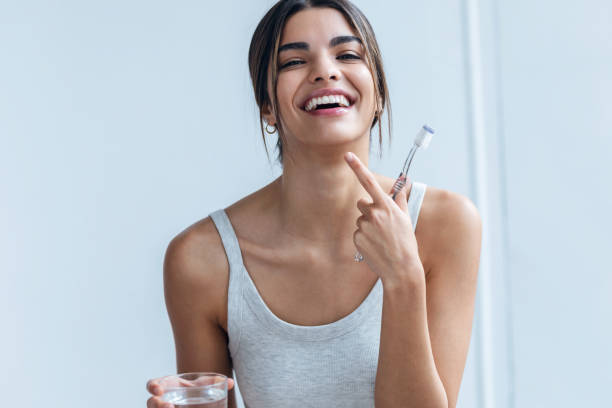 mulher jovem bonita escovando os dentes enquanto olha para a câmera no banheiro em casa. - toothbrush dental hygiene glass dental equipment - fotografias e filmes do acervo