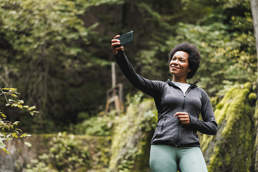 Mature African American woman making a video call while standing near the waterfall and enjoying the view during her hike in the mountains.