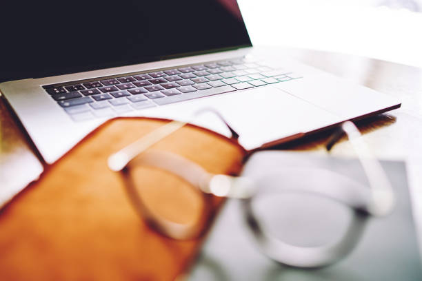 Closeup of modern laptop with black screen placed on wooden table near blurred notebook with leather cover and eyeglasses in daylight