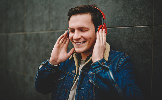 Happy male in casual clothes with toothy smile and closed eyes standing near dark wall while using headphones for listening to music