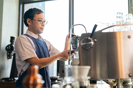 Frequent practice in expertise improve making coffee skill. Asia Japanese man or barista in apron standing at coffee shop counter bar and using coffee machine to make coffee under sunlight with smile. Enjoy work at restaurant. Food and drink business. Small business and working concept.