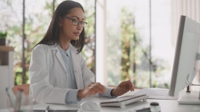 Young Female Doctor Wearing White Coat, Working on Personal Computer in Her Office. Beautiful Asian Medical Health Care Professional Working with Test Results, Patient Treatment Planning