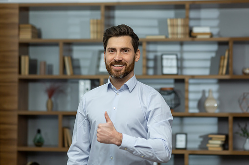 Successful businessman smiling and looking at camera, man shows thumbs up affirmatively and joyfully agrees with achievement, boss in shirt inside office at work.