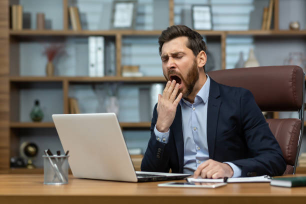 Mature man yawning inside classic office, senior businessman in workplace sleepless and overworked, boss in business suit sitting at desk, using laptop at work Mature man yawning inside classic office, senior businessman in workplace sleepless and overworked, boss in business suit sitting at desk, using laptop at work. lazy construction laborer stock pictures, royalty-free photos & images