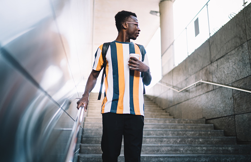 Pensive male tourist with caffeine beverage in hand standing at urban stairs and looking away pondering on solo travelling, African American hipster guy with takeaway coffee to go thinking in city