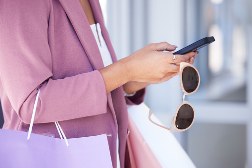 Woman, phone and shopping while holding sunglasses in hands and doing online shopping, ecommerce and banking. Female using mobile app for customer discount or coupon on commerce website at a mall