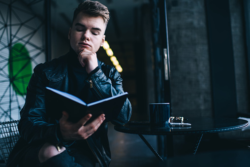 Serious young male in trendy leather jacket reading book hand at chin while enjoying cup of coffee in modern cafe