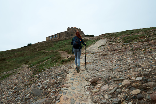 Low angle of female tourist with backpack and walking sticks ascending hill slope with old castle on gray day in countryside
