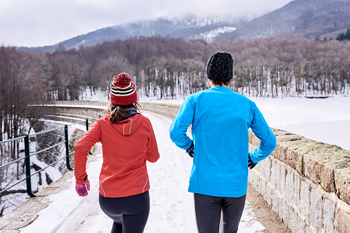 Back view of anonymous couple in sportswear and hat running on snowy bridge near forest and mountains during cardio training on winter day
