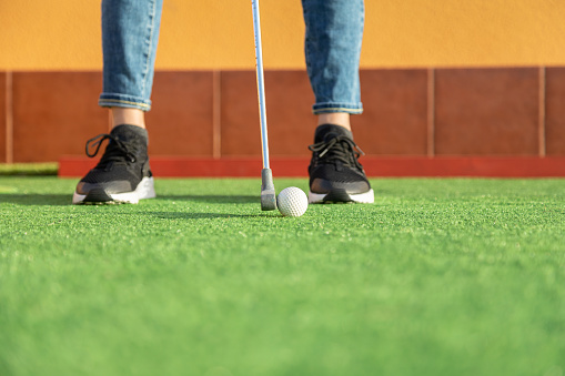 Close-up of unrecognizable woman hitting ball with golf club in mini golf and putting it in the hole