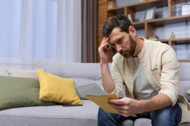 homem chateado em casa sentado no sofá lendo carta com notificação de más notícias na abertura do envelope, infeliz na sala de estar e triste - reading newspaper 30s adult - fotografias e filmes do acervo