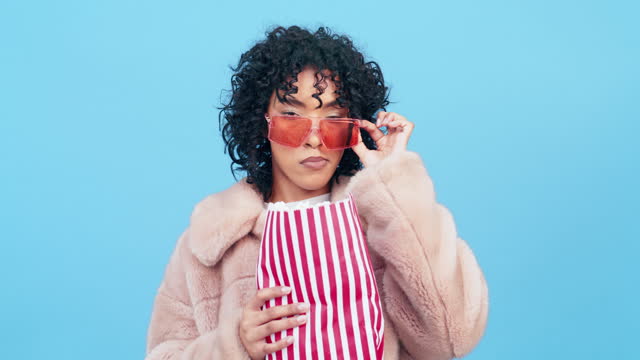 Fashion, popcorn and confidence with a black woman in studio on a blue background eating a snack. Food, glasses and style with an attractive young female enjoying popped corn at the movie or cinema