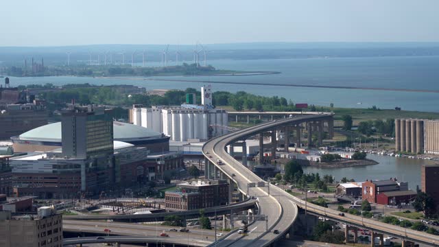 An aerial view of the city of Buffalo, New York and its infrastructure of bridges and buildings beside Lake Erie with some wind turbines in the background.