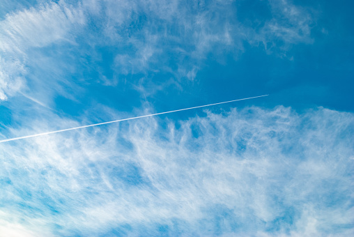 Cirrus clouds and long airplane trail row. Aero plane contrail in blue cloudy sky background. Horizontal line track from flying fast aeroplane in distance
