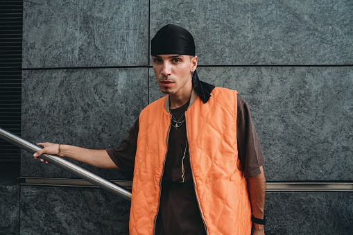 Portrait of a young adult man wearing cool urban clothes in the city. He's standing on a staircase against a grey wall.