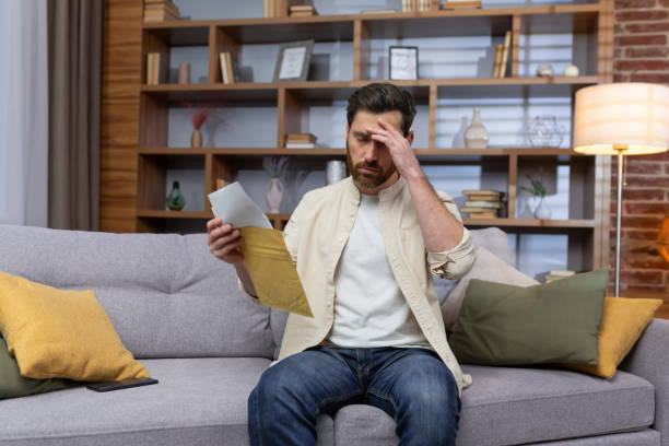 istock Upset man at home sitting on sofa reading letter with bad news notification in envelope opening, unhappy in living room and sad 1473957795